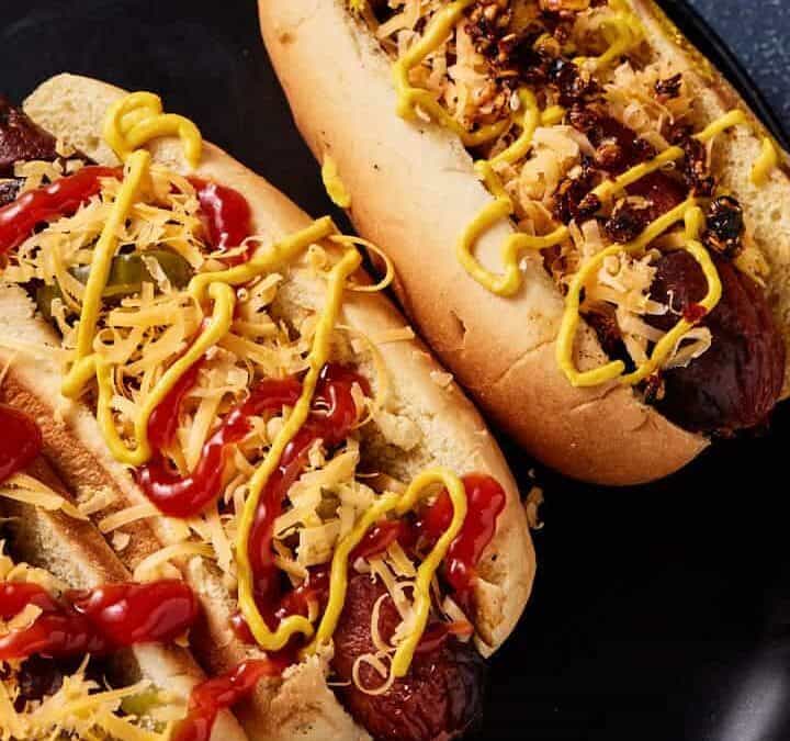 Two grilled hot dogs in buns, topped with shredded cheese, ketchup, mustard, and relish, served on a dark plate with a dark background.