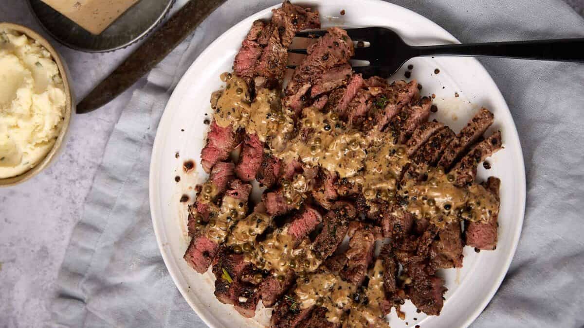 Sliced steak au poivre topped with creamy peppercorn sauce on a white plate, with a black fork, vintage knife, mashed potatoes in a bowl, and extra sauce au poivre in a glass nearby, all set on a light gray cloth.