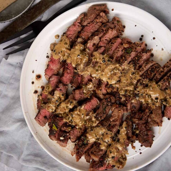 Sliced steak au poivre topped with peppercorn sauce on a white plate, accompanied by a fork and knife. In the background, there’s a small bowl of mashed potatoes and a block of sauce, all set on a gray cloth.