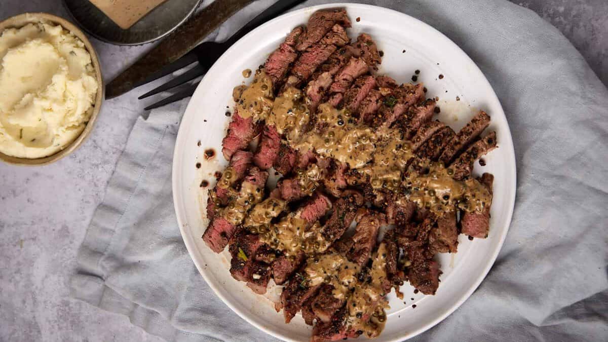 Sliced steak au poivre topped with peppercorn sauce on a white plate, accompanied by a fork and knife. In the background, there’s a small bowl of mashed potatoes and a block of sauce, all set on a gray cloth.
