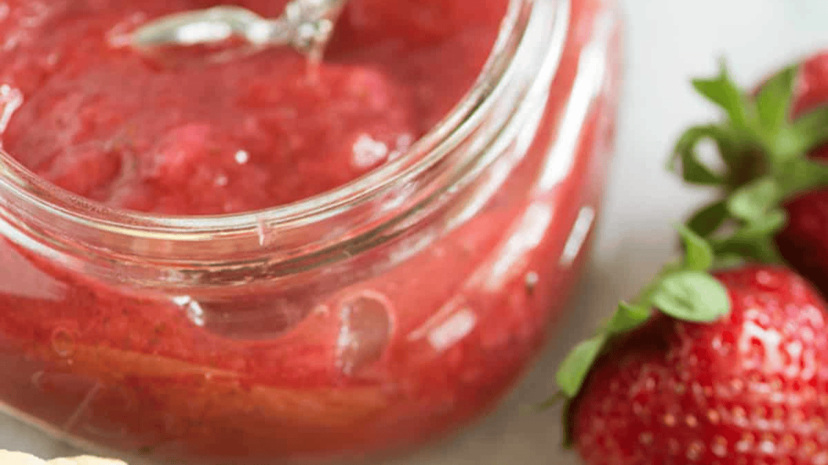 A close-up of a glass jar filled with strawberry jam, with fresh strawberries placed nearby on a white surface.