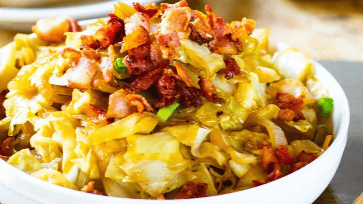 A bowl of cooked cabbage mixed with crispy bacon pieces, garnished with green onions, served on a light-colored plate.