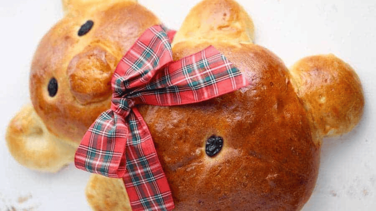 A loaf of bread shaped like a bear, decorated with a red and green plaid ribbon around its neck and raisins for eyes and nose, placed on a white surface.