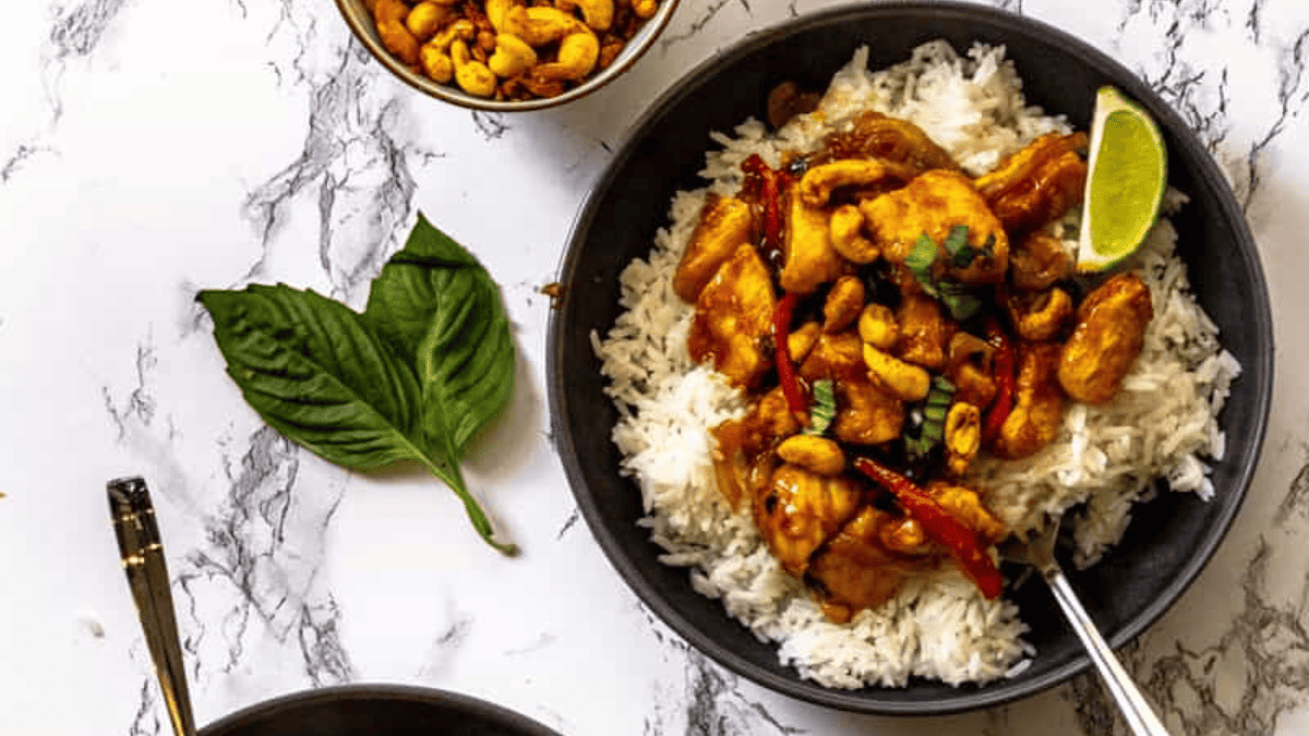 A bowl of white rice topped with stir-fried chicken, cashews, red chilies, and a lime wedge sits on a marble surface. A spoon, fresh basil leaves, and a small bowl of cashews are nearby.