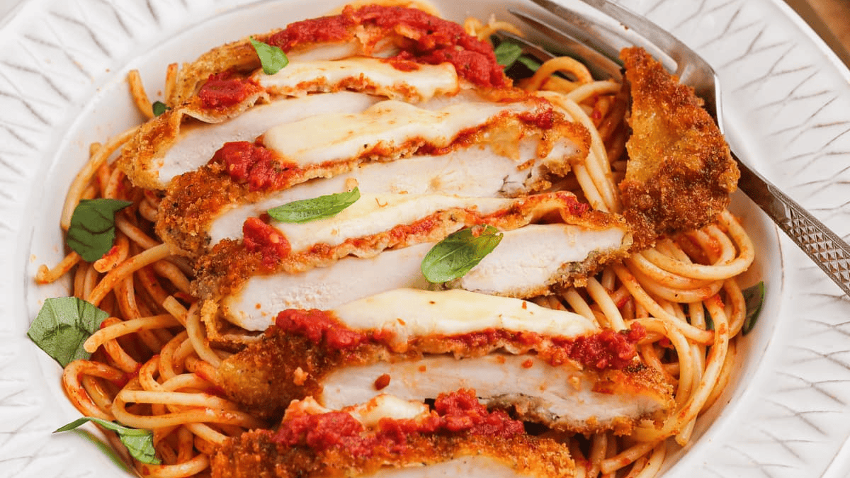 A plate of spaghetti topped with sliced breaded chicken, melted cheese, marinara sauce, and fresh basil, with a fork on the side.