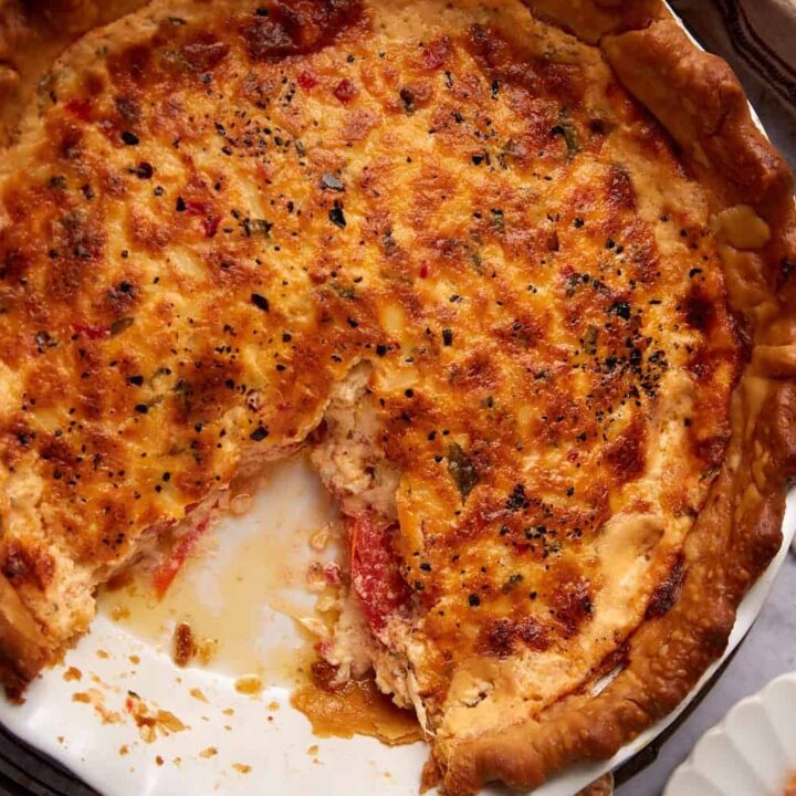 A baked savory pimento cheese tomato pie with a golden, flaky crust sits on a round pan, one slice removed and placed on a white plate beside it. A beige napkin is nearby on a textured surface.