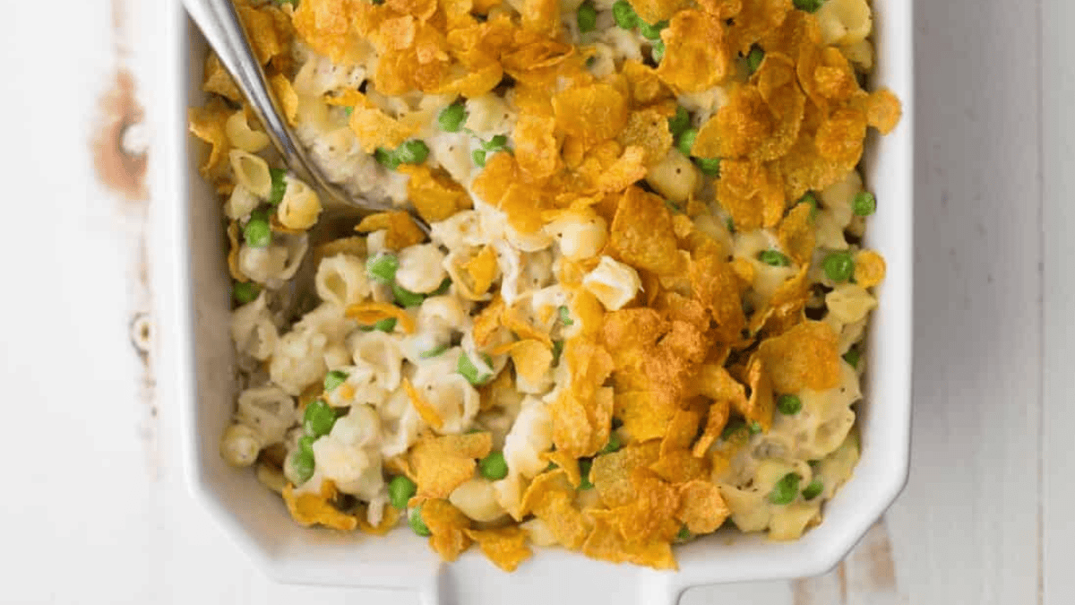 A close-up of a casserole dish filled with creamy pasta, green peas, and topped with golden, crispy cornflake crumbs. A spoon is partially visible in the dish.