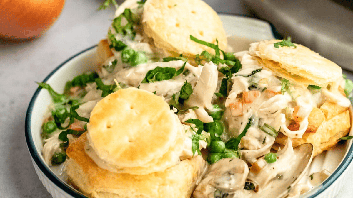 A plate of creamy chicken and vegetables, including peas and carrots, served over flaky biscuit halves and garnished with chopped fresh herbs.