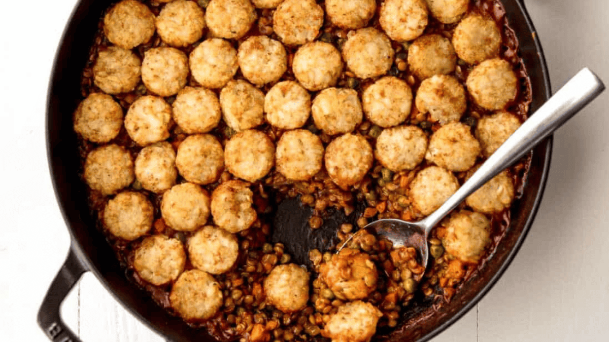 A cast iron skillet filled with tater tot casserole; golden tater tots arranged on top, with a section scooped out revealing a filling of peas, carrots, corn, and ground meat. A metal serving spoon rests in the casserole.