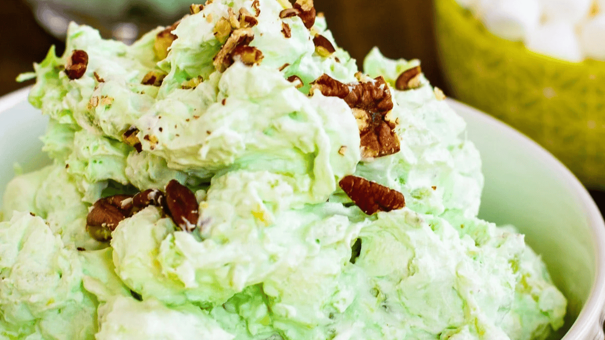 A white bowl filled with a fluffy, light green salad topped with chopped pecans. In the background, another bowl with white and yellow ingredients is slightly visible.