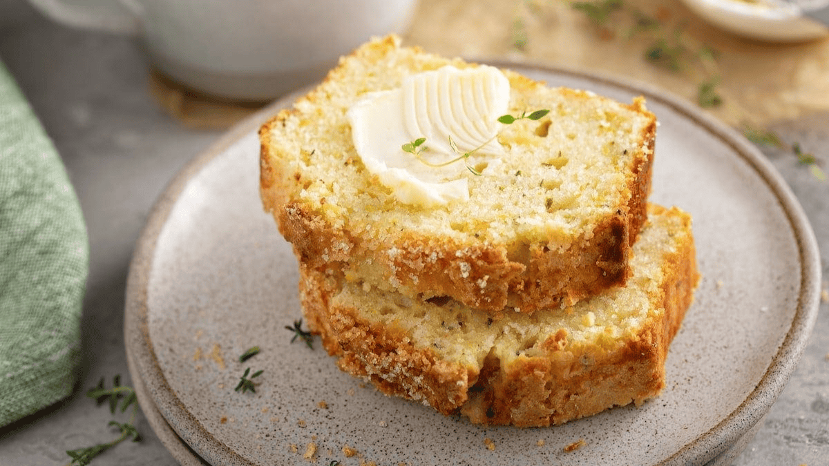 Two slices of zucchini bread stacked on a plate, with a pat of butter melting on top. Small sprigs of fresh herbs are sprinkled around, and a green napkin is visible in the background.