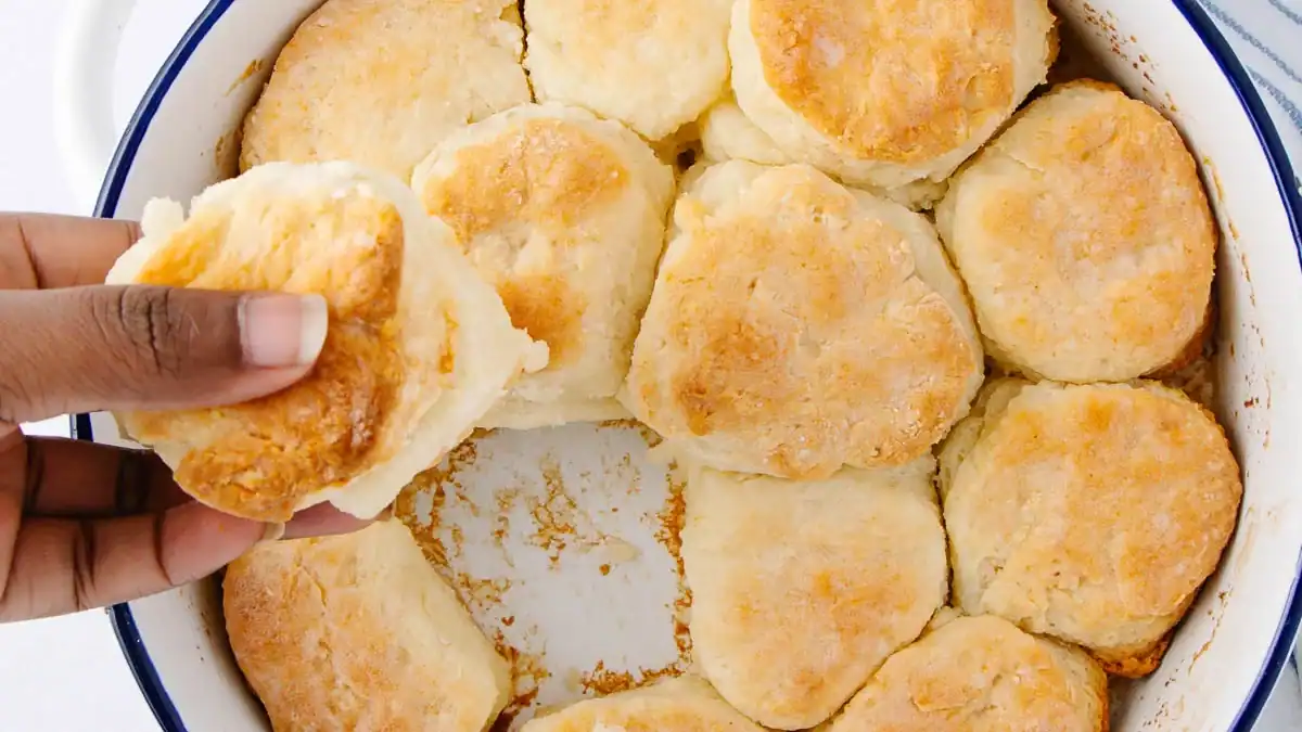 A hand lifts a golden, fluffy biscuit from a round white baking dish filled with freshly baked biscuits. A striped kitchen towel lies nearby on the white surface.