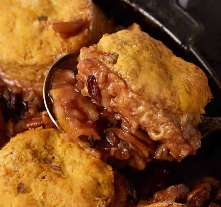 A skillet filled with Apple Cobbler with Sweet Potato Biscuits atop a caramelized filling, being served with a spoon. A small white pitcher sits in the background on a dark coaster.