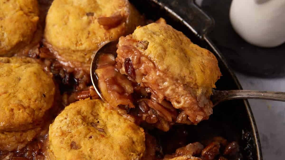 A skillet filled with Apple Cobbler with Sweet Potato Biscuits atop a caramelized filling, being served with a spoon. A small white pitcher sits in the background on a dark coaster.