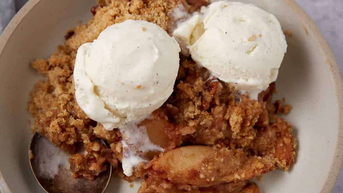 A bowl of apple crisp with oatmeal topping, topped with two scoops of vanilla ice cream, with a spoon resting in the bowl. The dessert sits on a light gray surface with a cloth napkin nearby.