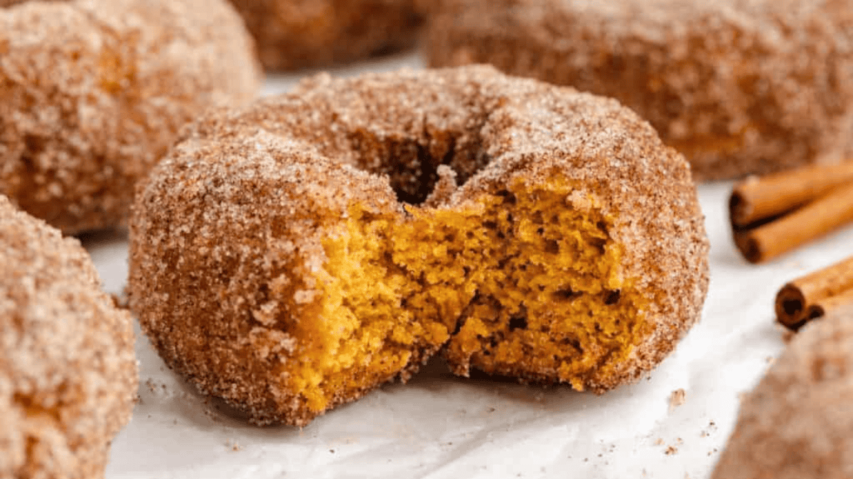 A close-up of a cinnamon sugar-coated donut with a soft, moist interior, split in half to show its texture. Other donuts and cinnamon sticks are visible in the background.