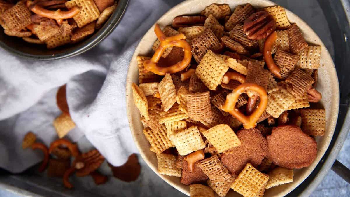 A bowl filled with bold Chex Mix—Chex cereal, pretzels, bagel chips, and pecans—sits on a tray next to another bowl of the same snack mix, with a light cloth napkin nearby, perfect for sharing your favorite Chex Mix recipe oven creation.