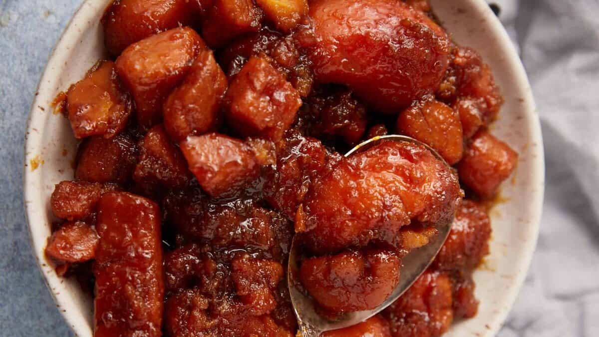 A bowl filled with southern candied yams and sweet potatoes, covered in a shiny brown sugar syrup, with a spoon scooping some out.