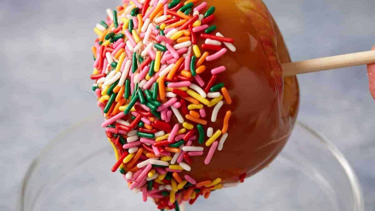 A caramel apple coated with colorful rainbow sprinkles is held by a wooden stick above a glass bowl—celebrate apple harvest season with these comforting classics.
