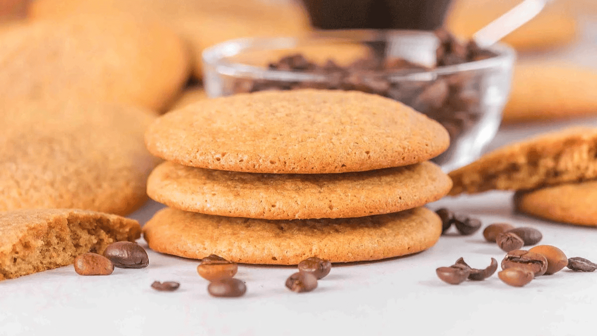 Three golden-brown cookies are stacked, surrounded by coffee beans and a bowl of beans in the background, with a broken cookie to the side&mdash;perfect for those exploring creative coffee recipes beyond the mug.