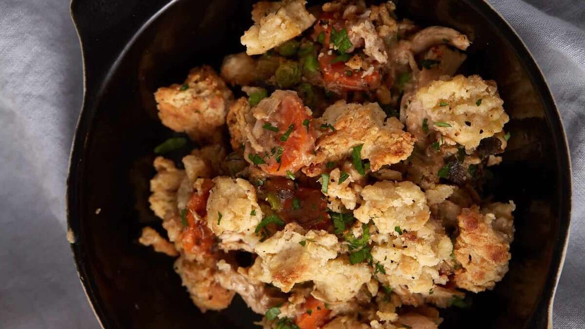 A black bowl is filled with a savory mixture of baked chicken, cauliflower, and carrots, garnished with fresh green herbs. The dish appears to be seasoned with spices and roasted to a golden brown. It is set on a light gray cloth.