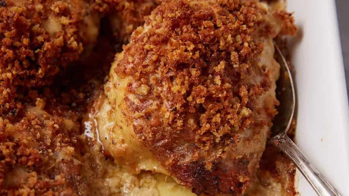 Golden-brown, crumb-topped oven baked chicken thighs arranged in a white baking dish, with a spoon lifting one piece to reveal melted cheese and savory juices underneath.