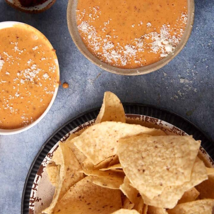 A plate of tortilla chips sits next to several bowls filled with creamy orange Chorizo Queso Dip, some sprinkled with grated cheese, on a blue surface.