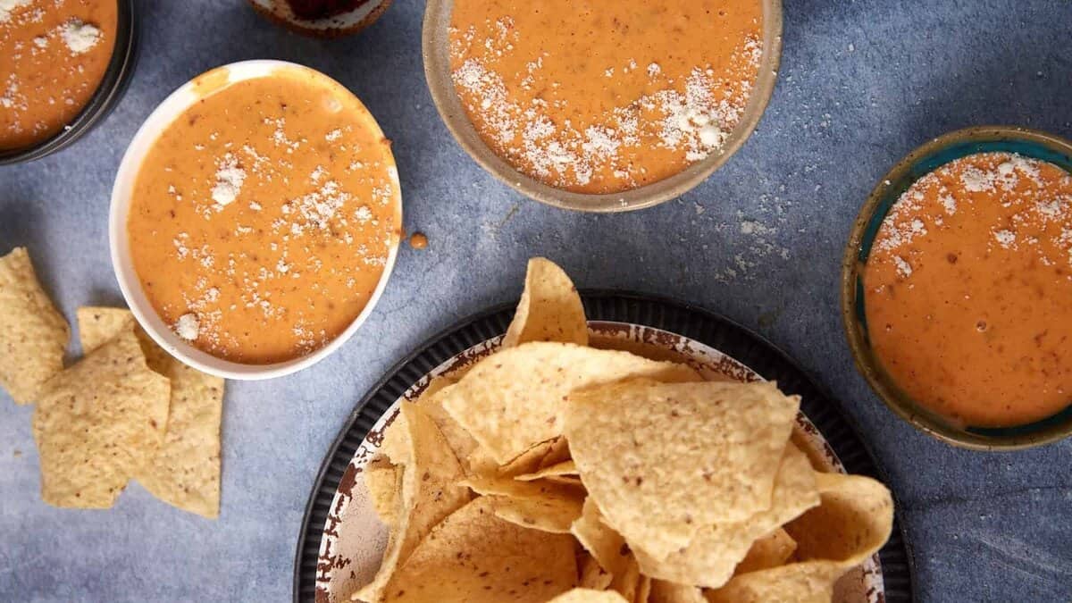 A plate of tortilla chips sits next to several bowls filled with creamy orange Chorizo Queso Dip, some sprinkled with grated cheese, on a blue surface.