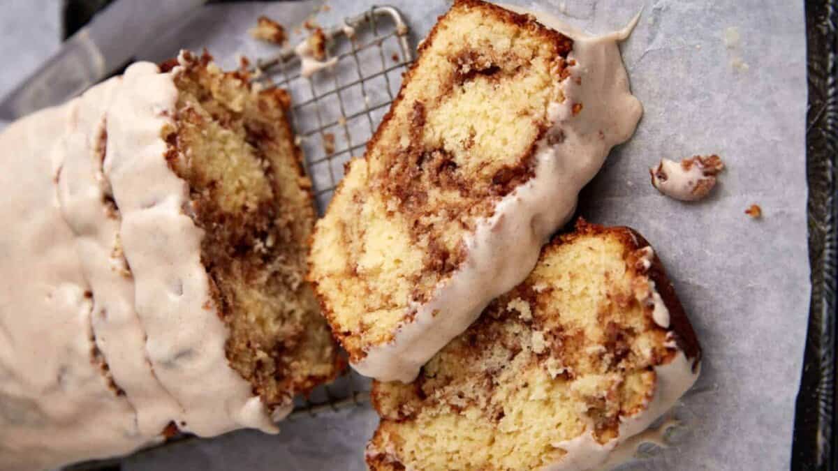 Two slices of glazed cinnamon swirl bread lie on parchment paper next to the loaf, showing a cinnamon-sugar spiral inside and a thick layer of icing on top. A cooling rack is partially visible beneath the bread.