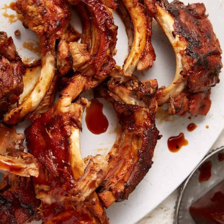 A white plate is filled with juicy, Coca Cola ribs covered in a rich, sticky sauce. A fork is lifting a piece of rib from the plate. A small metal dish of extra barbecue sauce is in the background. The surface underneath is light-colored.