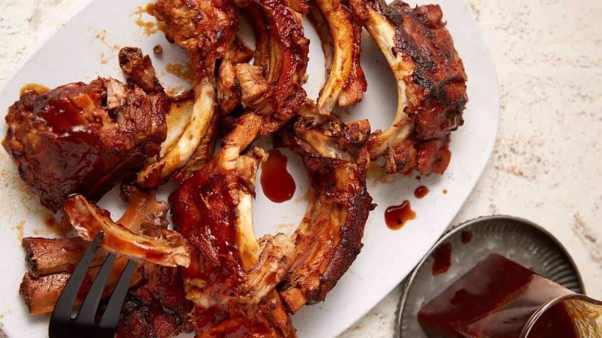 A white plate is filled with juicy, Coca Cola ribs covered in a rich, sticky sauce. A fork is lifting a piece of rib from the plate. A small metal dish of extra barbecue sauce is in the background. The surface underneath is light-colored.