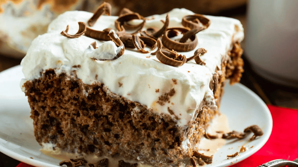 A slice of chocolate cake topped with whipped cream and chocolate curls sits on a white plate, perfect for pairing with creative coffee recipes beyond the mug. The moist cake and creamy topping are complemented by scattered chocolate shavings.