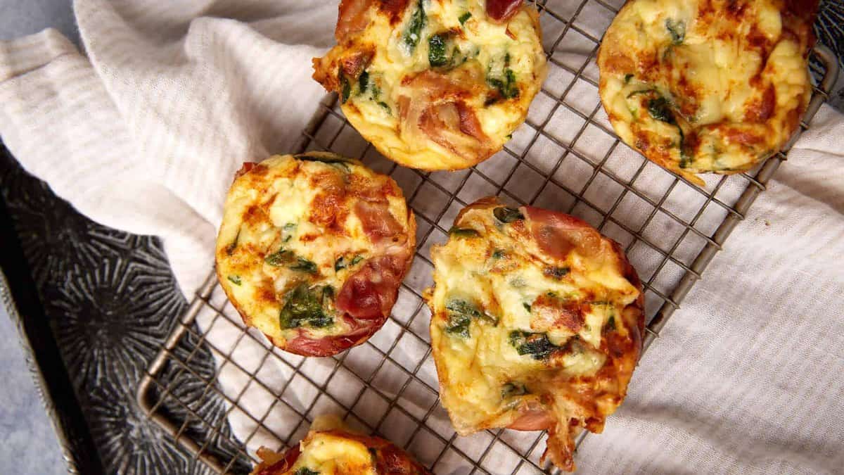 Five mini Prosciutto Egg Cups with spinach and ham sit on a cooling rack over a white cloth, showcasing a golden, crispy top and a fluffy interior with visible pieces of ham and green vegetables.