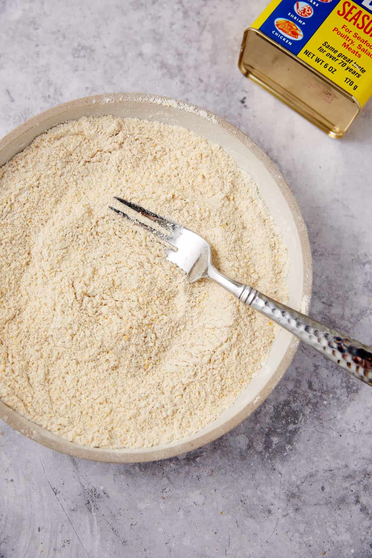 A bowl filled with a light yellow, fine powder mixture and a fork resting inside; next to the bowl is a rectangular metal tin labeled “Seasoned Flour.”.