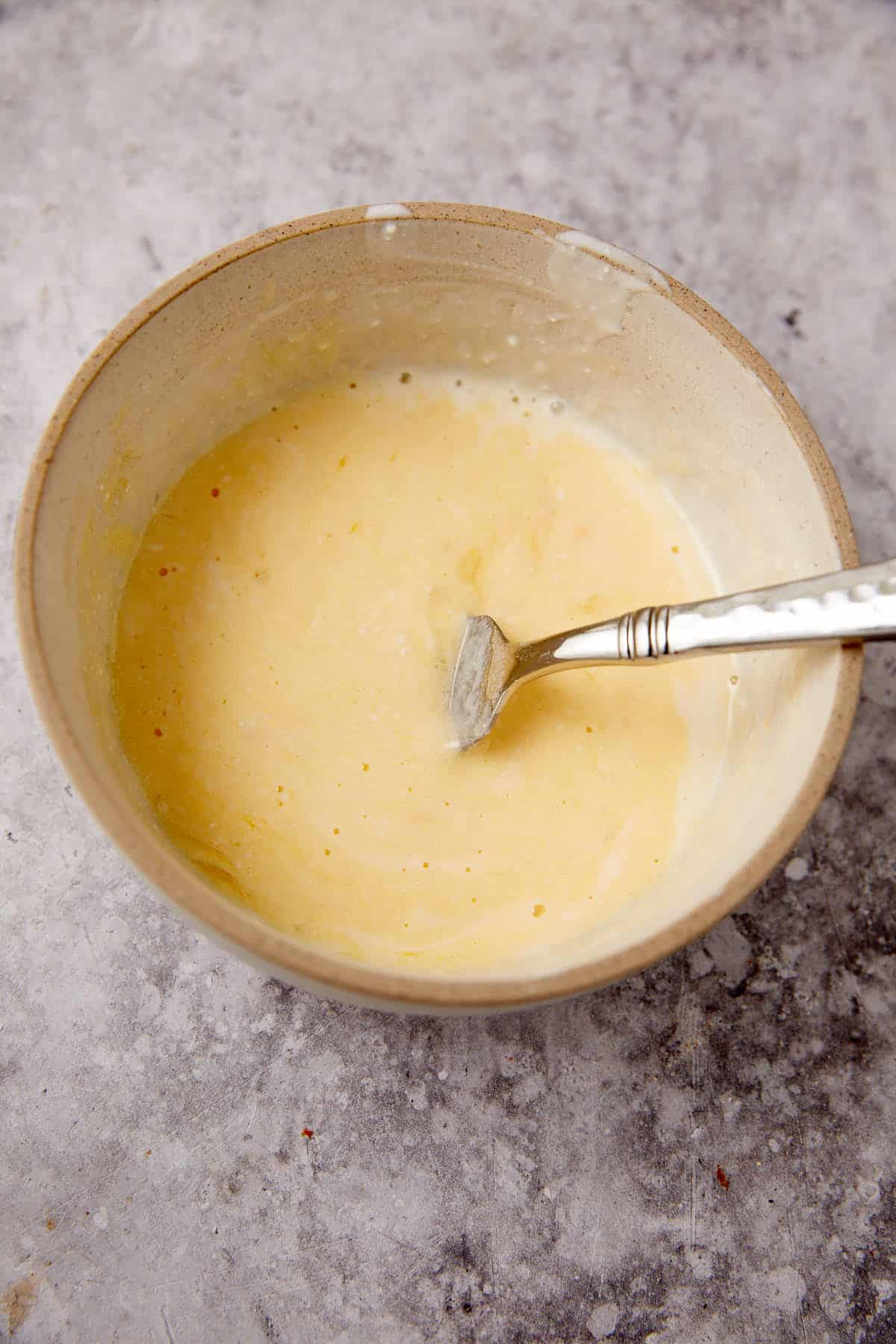 A bowl filled with a light yellow batter, with a fork resting inside. The bowl sits on a gray mottled surface.