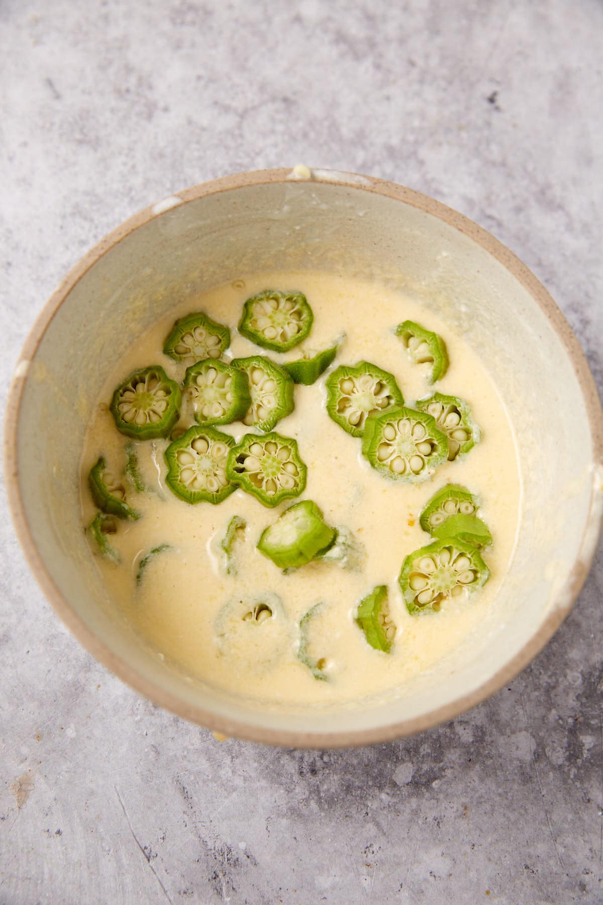 A bowl filled with a creamy yellow batter and slices of fresh green okra on top, placed on a light gray textured surface.