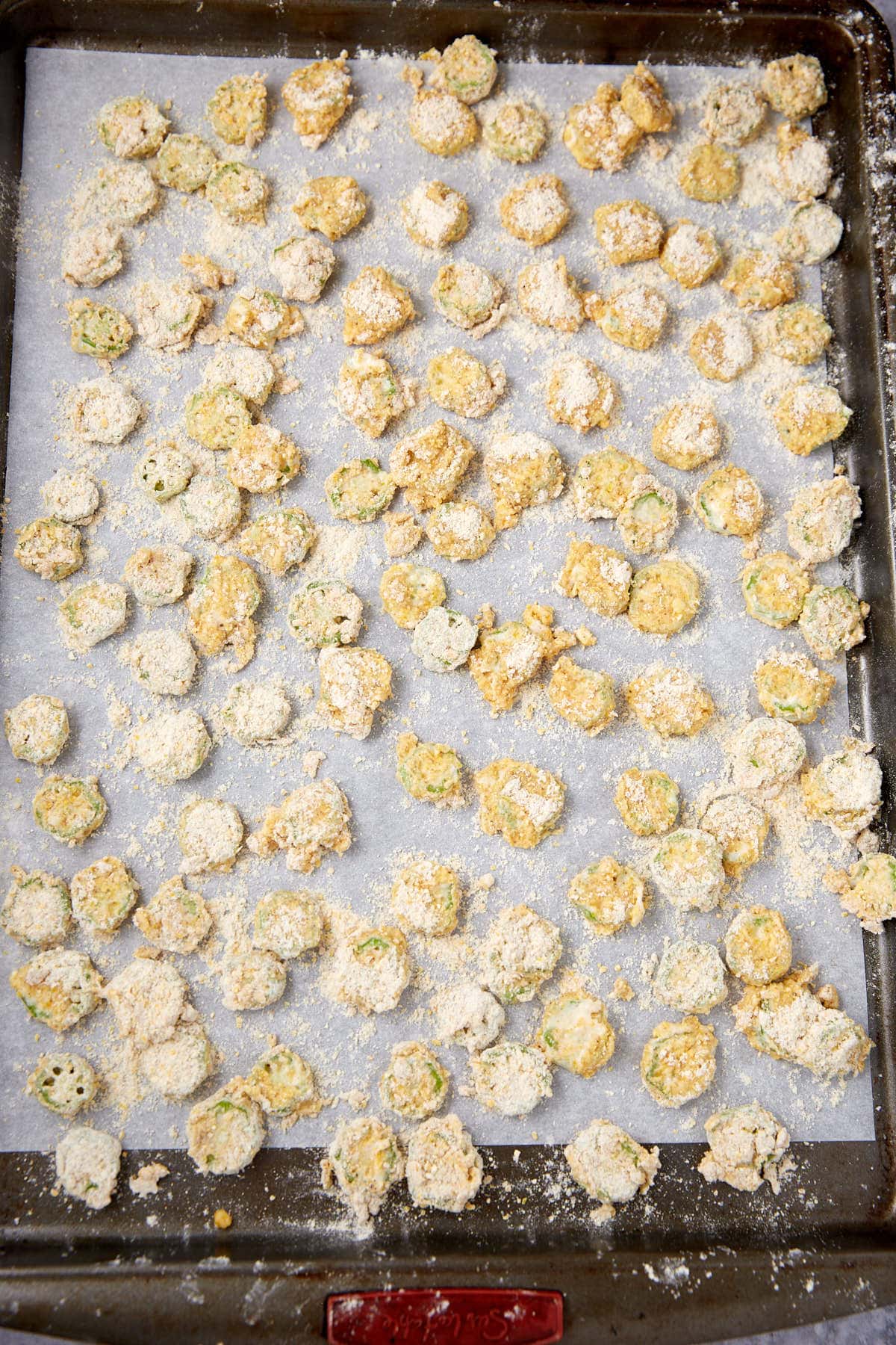 A baking sheet lined with parchment paper, covered with evenly spaced pieces of sliced okra coated in a light layer of flour and breading, ready to be baked or fried.