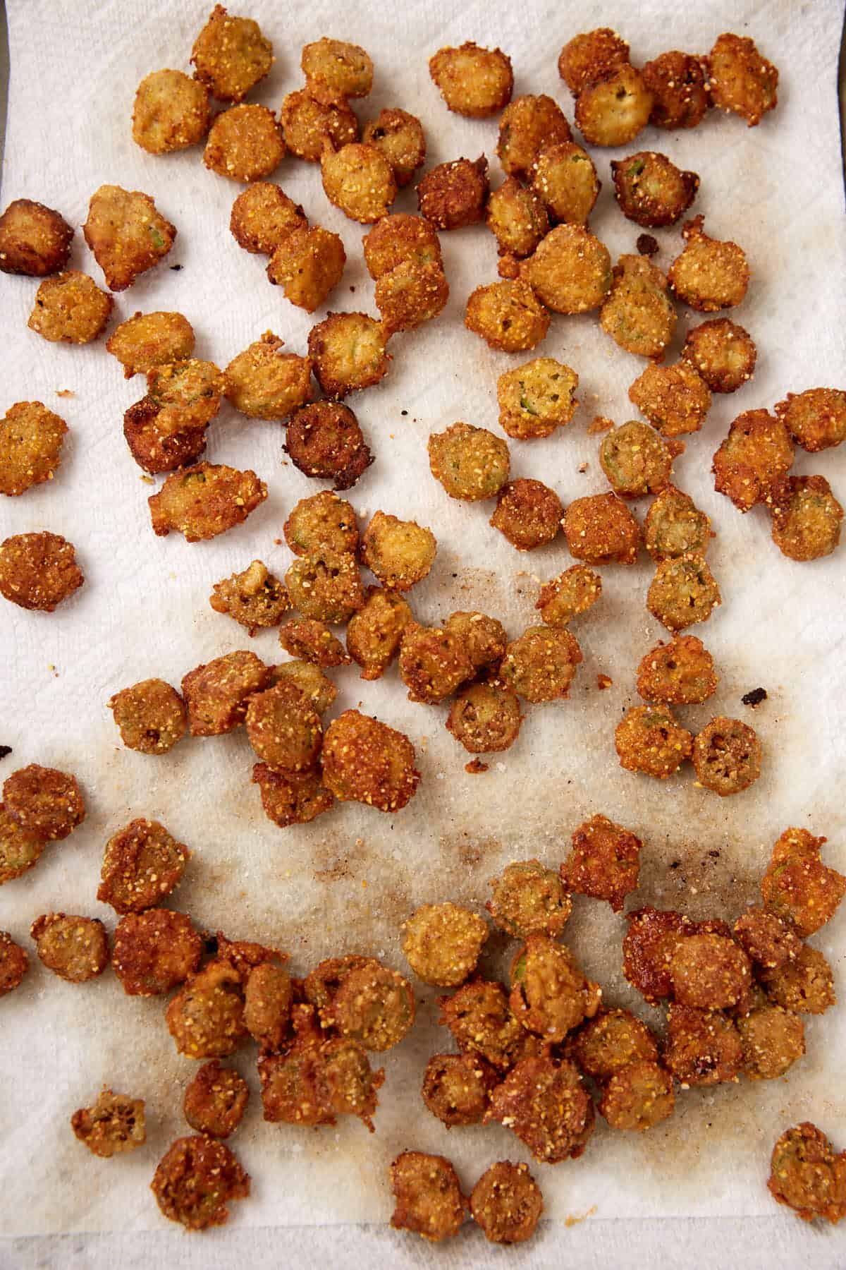 Golden brown pieces of fried okra are scattered on a white paper towel, which is absorbing excess oil from the fried food.