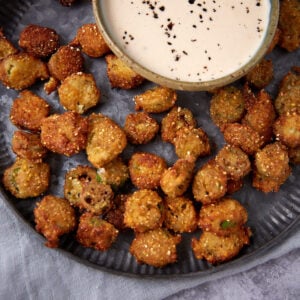 A plate of crispy fried okra pieces served with a bowl of creamy dipping sauce topped with cracked black pepper.