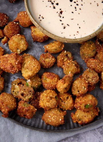 A plate of crispy fried okra pieces served with a bowl of creamy dipping sauce topped with cracked black pepper.