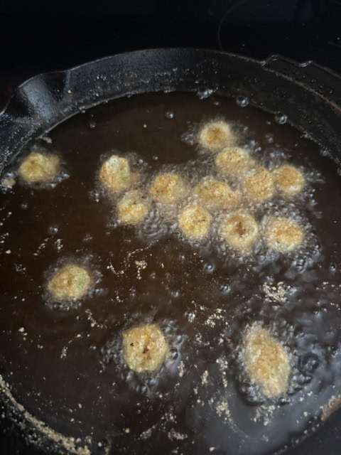 Small, round pieces of battered food are being deep fried in hot oil in a black cast iron skillet, with bubbles forming around each piece as they cook.