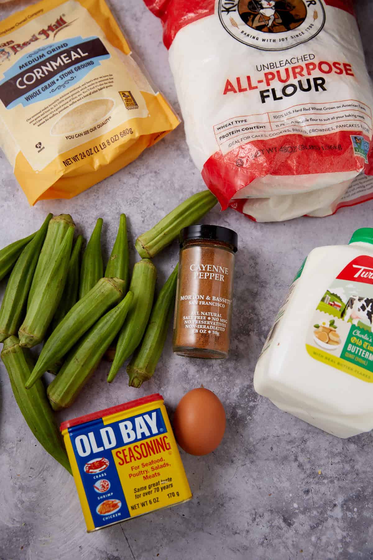 A flat lay of cooking ingredients: a bag of cornmeal, a bag of all-purpose flour, fresh okra pods, a container of Old Bay seasoning, a bottle of cayenne pepper, a carton of buttermilk, and a brown egg on a gray surface.