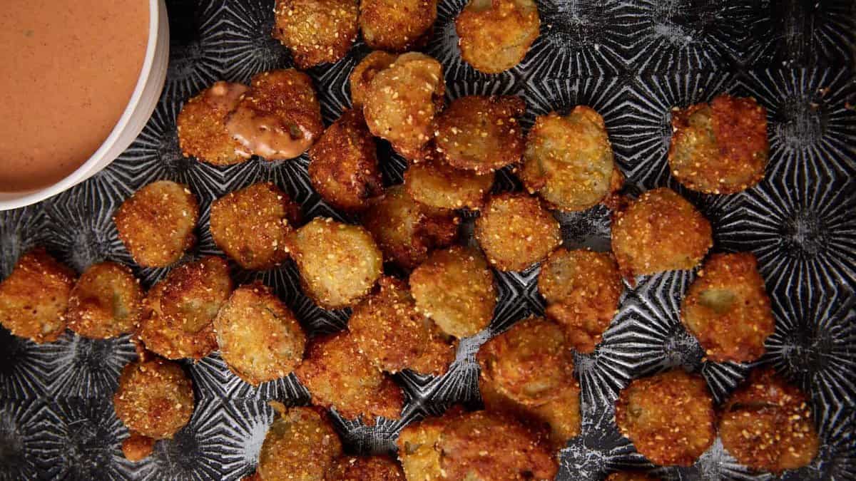 A batch of golden-brown Southern Fried Pickles is scattered on a dark textured tray, with a small bowl of creamy dipping sauce in the corner.