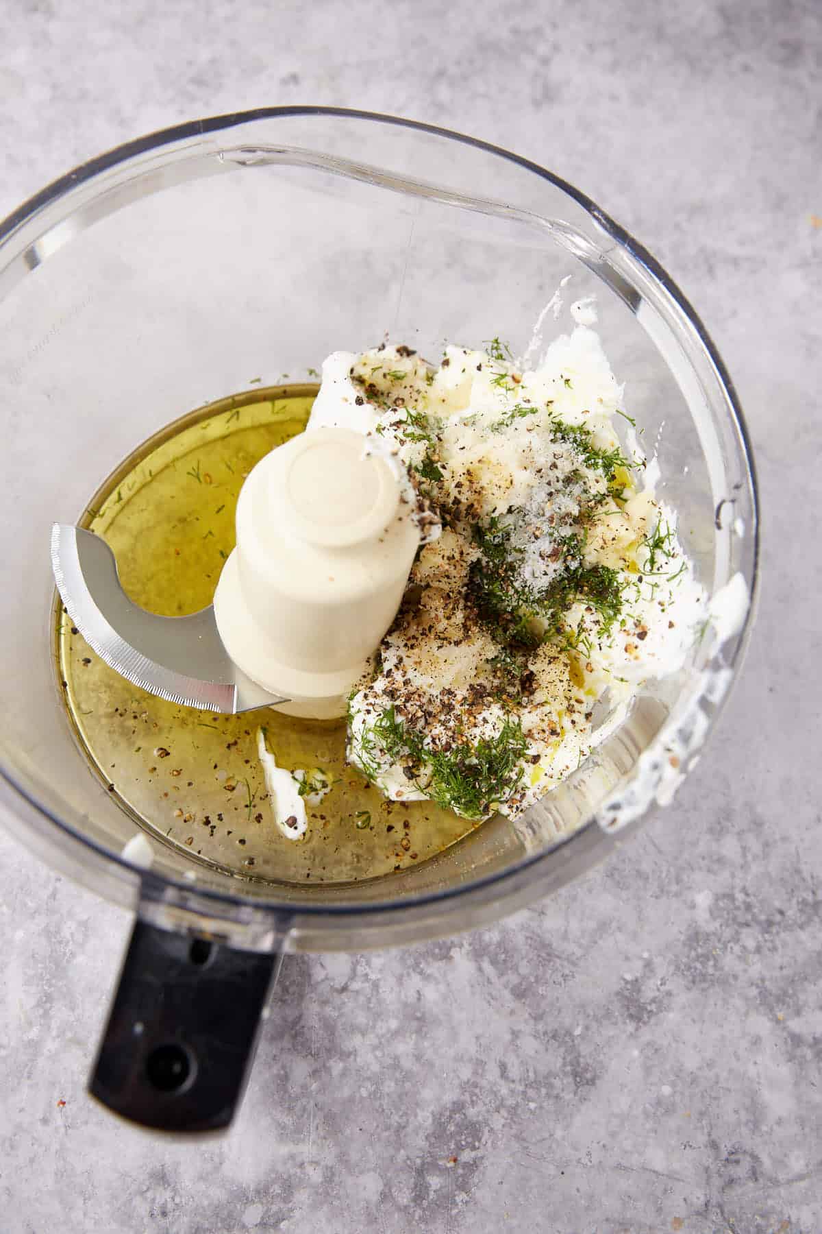 A food processor bowl containing olive oil, cream cheese, herbs, and black pepper, ready to be blended, on a gray countertop.