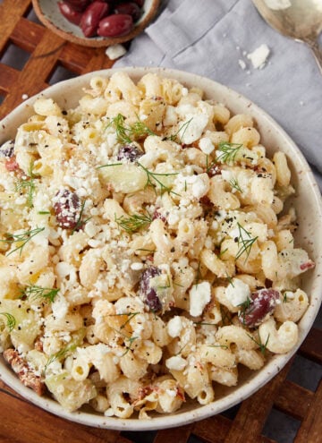 A bowl of pasta salad with rotini, crumbled feta cheese, olives, cucumber, and fresh dill sits on a wooden surface next to a spoon and a small dish of olives.