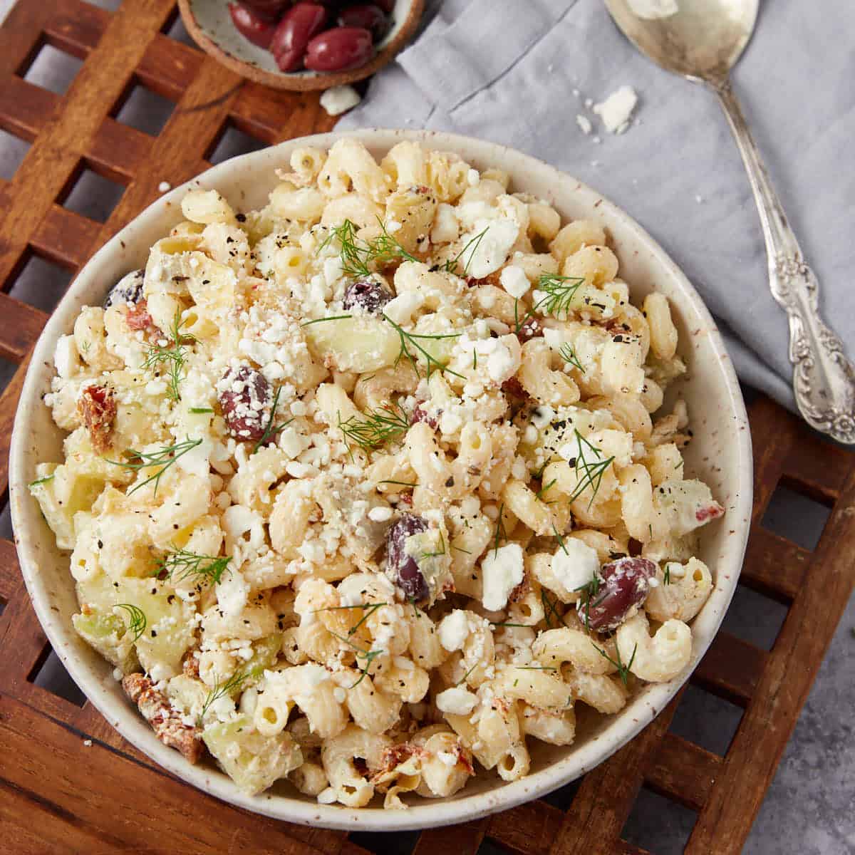 A bowl of pasta salad with rotini, crumbled feta cheese, olives, cucumber, and fresh dill sits on a wooden surface next to a spoon and a small dish of olives.