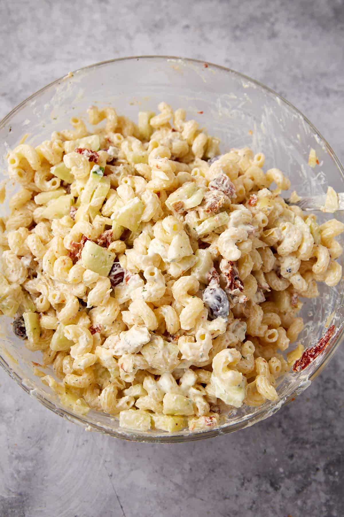 A glass bowl filled with creamy pasta salad, featuring elbow macaroni, chopped celery, raisins, and sun-dried tomatoes, all mixed together on a textured gray surface.