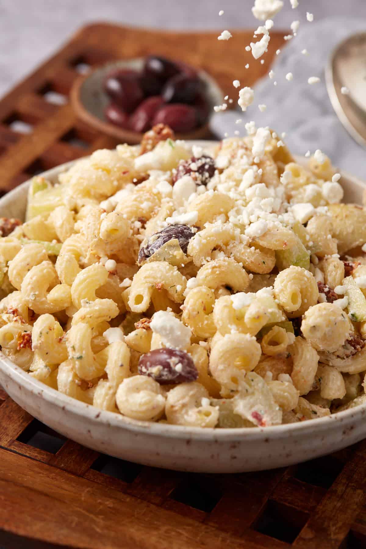 A bowl of creamy pasta salad with olives and crumbled feta cheese being sprinkled on top, placed on a wooden tray with a small dish of olives in the background.