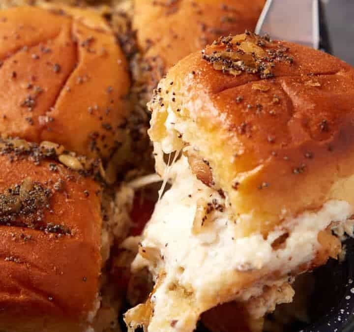 A hand uses a spatula to lift a cheesy Hot Brown Slider from a pan filled with golden, seasoned slider sandwiches.