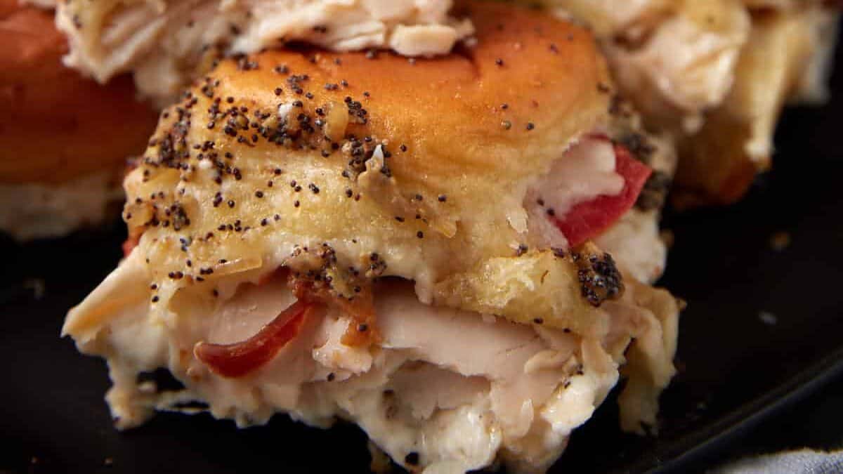 A close-up of stacked Hot Brown Sliders filled with turkey, cheese, and tomato, topped with a poppy seed seasoning mix, served on a dark plate. A blurred plate of tomato slices is visible in the background.